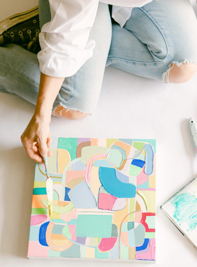 photo of Amanda Petro kneeling on floor working on a painting with geometric shapes and a mix of bright and pastel colors