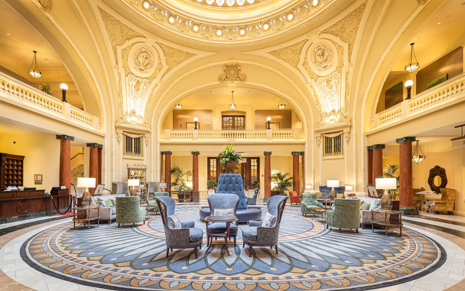 A grand, symmetrical hotel lobby with ornate cream and gold architectural details, featuring multiple blue upholstered armchairs, green side tables, and a large circular patterned rug.