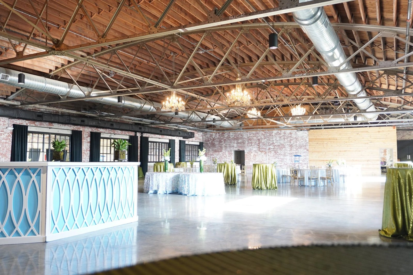 A large indoor event space with exposed wood ceiling beams, metal ducts, and hanging lights. The room contains several round tables with white tablecloths and draped table covers in olive green, plus a light blue decorative barrier in the foreground.