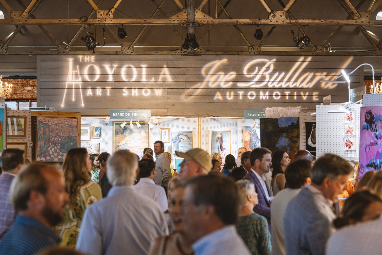 A crowded indoor event space with many people in the foreground and a large illuminated sign reading 'Theoyola Art Show' and 'Joe Bullard Automotive' above a booth displaying framed artwork.