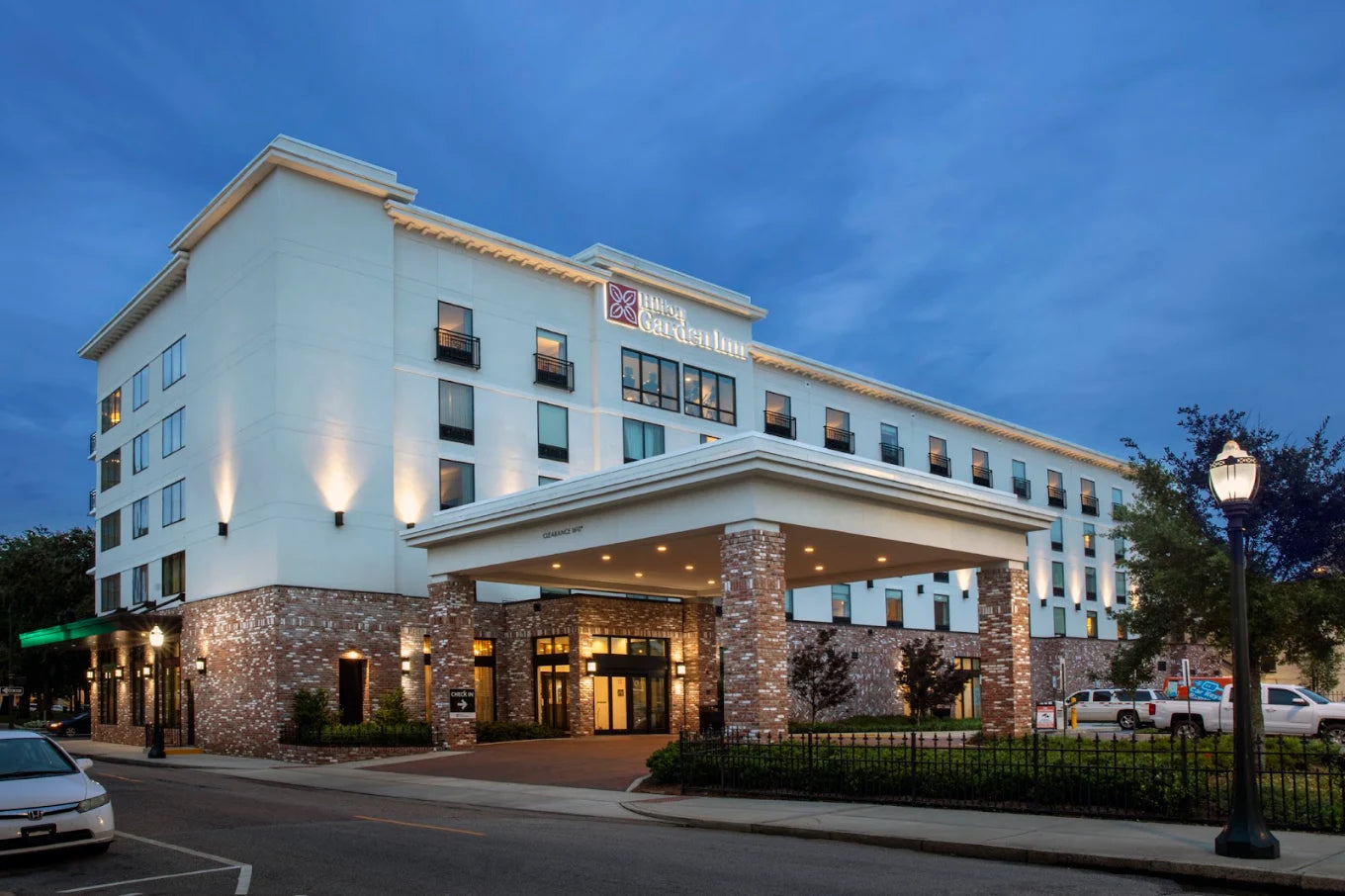 Hotel Granddellans hotel exterior at dusk, showing a multi-story white building with stone base columns, illuminated windows, a covered entrance, parked cars, and a street lamp in the foreground.