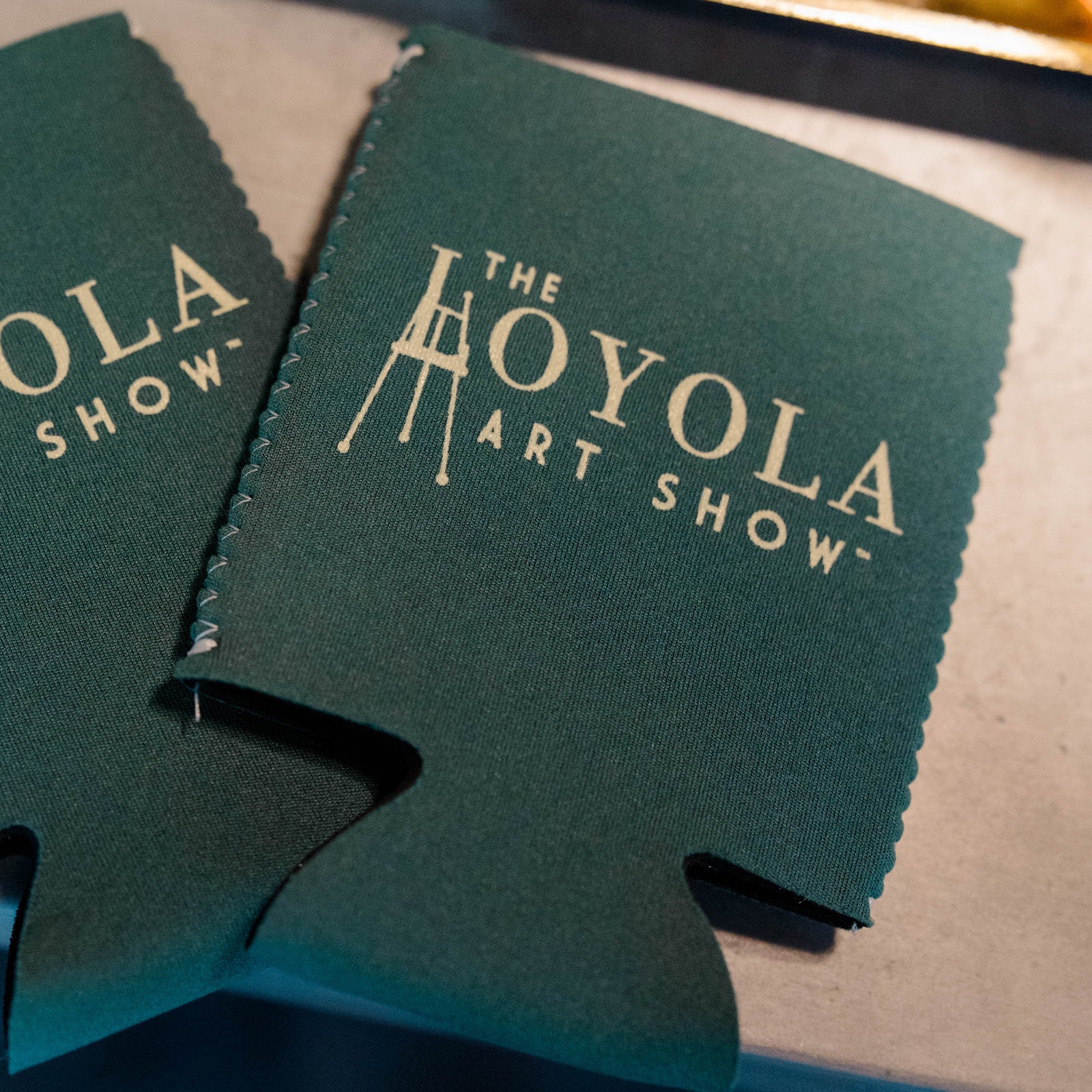 Two green can koozies with 'The Loyola Art Show' logo on a metallic surface.