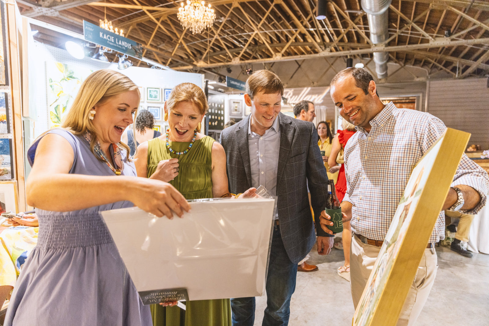 Four people gathered around a large white board or canvas at what appears to be a craft fair or market, with a man in a plaid shirt holding a wooden sign in the foreground. The scene is indoors with a visible ceiling and other booths in the background.