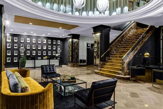 the admiral hotel A luxury hotel lobby featuring a grand staircase with gold-toned steps and a dark wood banister, modern black leather armchairs, a mustard yellow velvet sofa, and a glass coffee table on a polished stone floor.